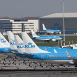 Six KLM blue and white planes are in a line on the tarmac with their tails as the centre of the image.