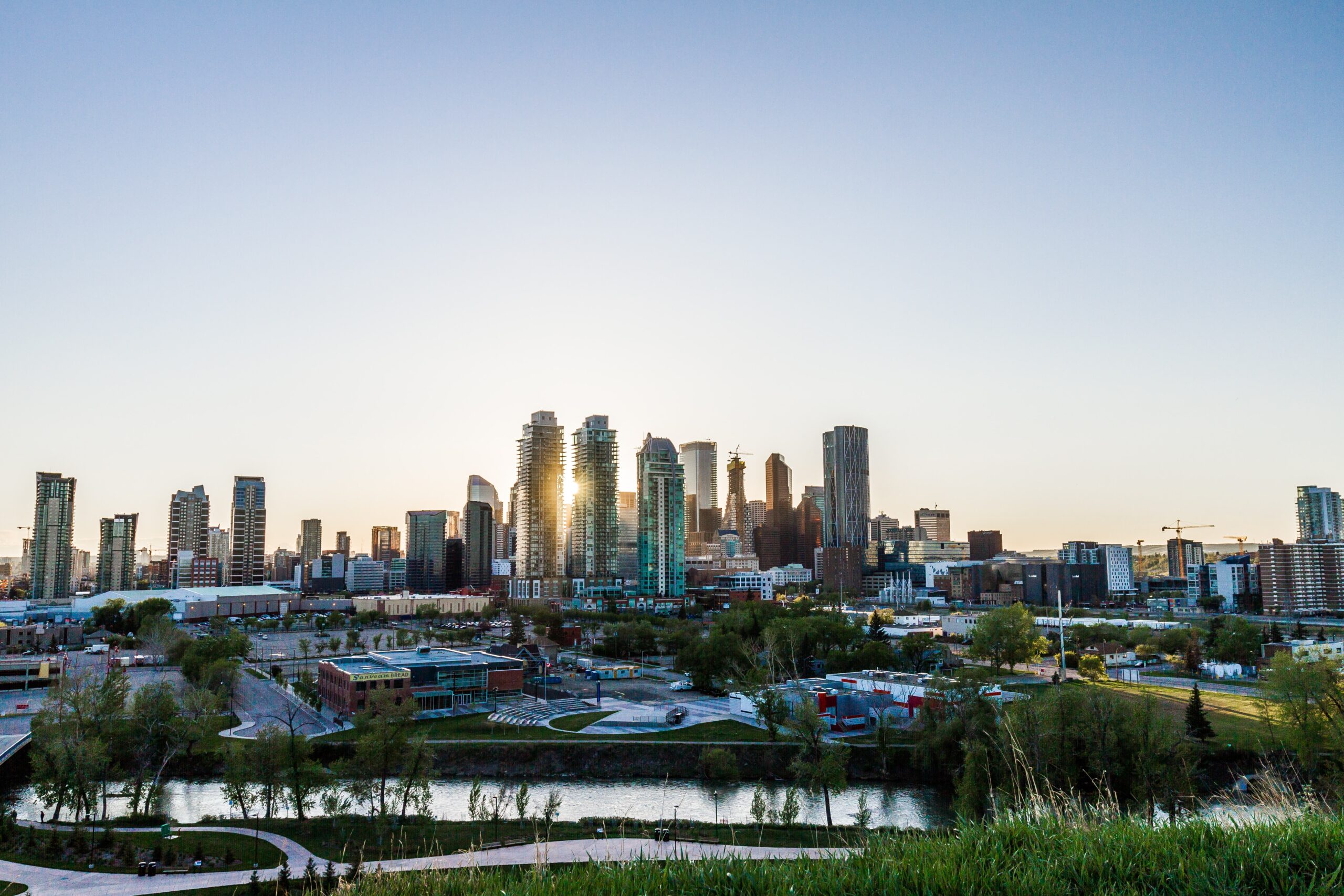 A panoramic view of a city skyline during sunset, featuring numerous modern high-rise buildings with glass facades. The sun is partially hidden behind the tallest buildings, casting a warm glow. In the foreground, there is a river with green spaces and pathways along its banks.