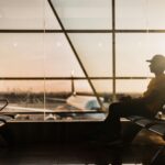A person wearing a cap and a yellow shirt sits alone in an airport terminal, silhouetted against the large windows as the sun sets. An airplane is visible outside on the tarmac.