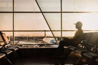 A person wearing a cap and a yellow shirt sits alone in an airport terminal, silhouetted against the large windows as the sun sets. An airplane is visible outside on the tarmac.