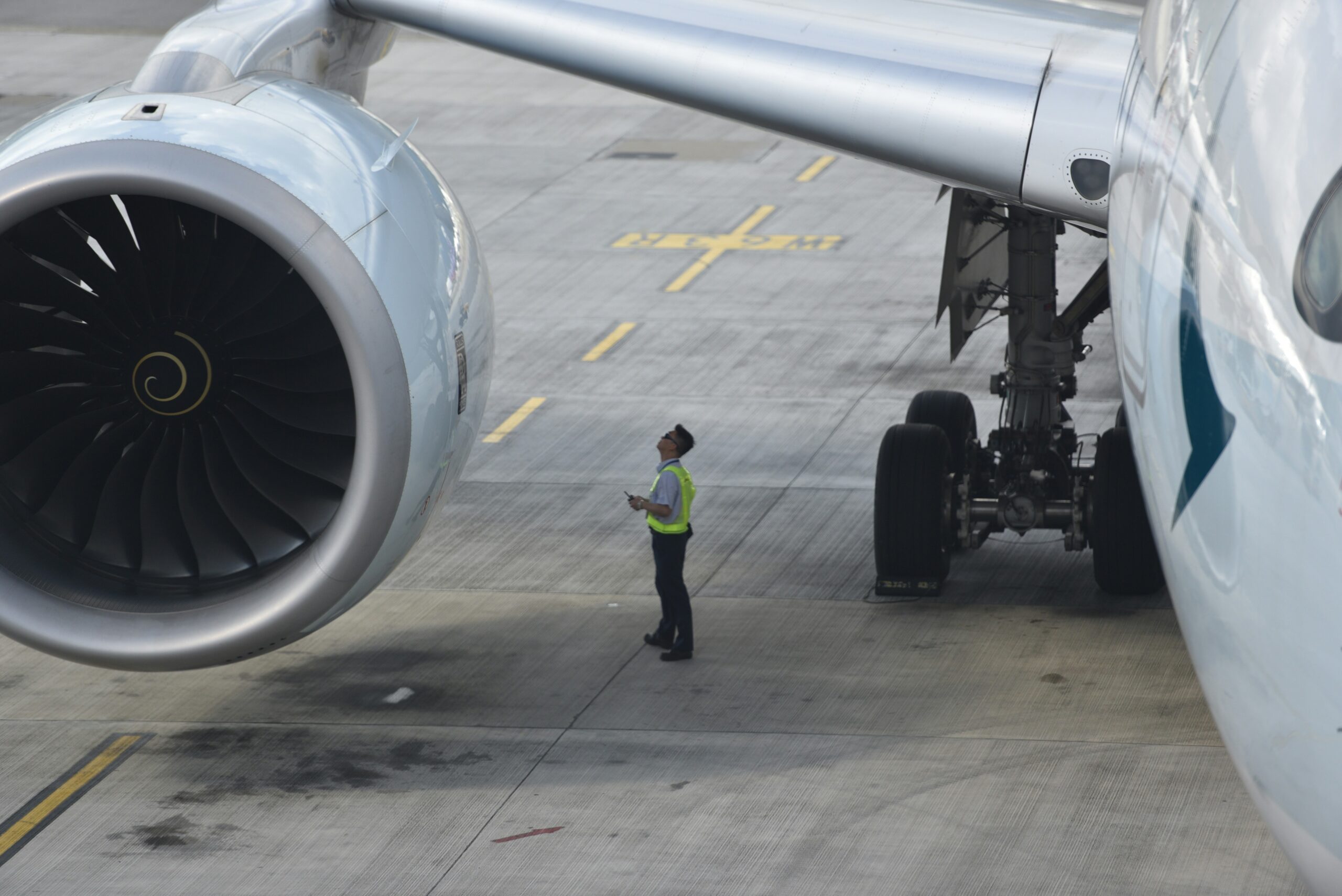  A ground crew member in a high-visibility vest is inspecting the engine of a large commercial airplane on an airport tarmac. The image shows the aircraft's jet engine and landing gear, with the crew member standing between them, looking up at the engine.