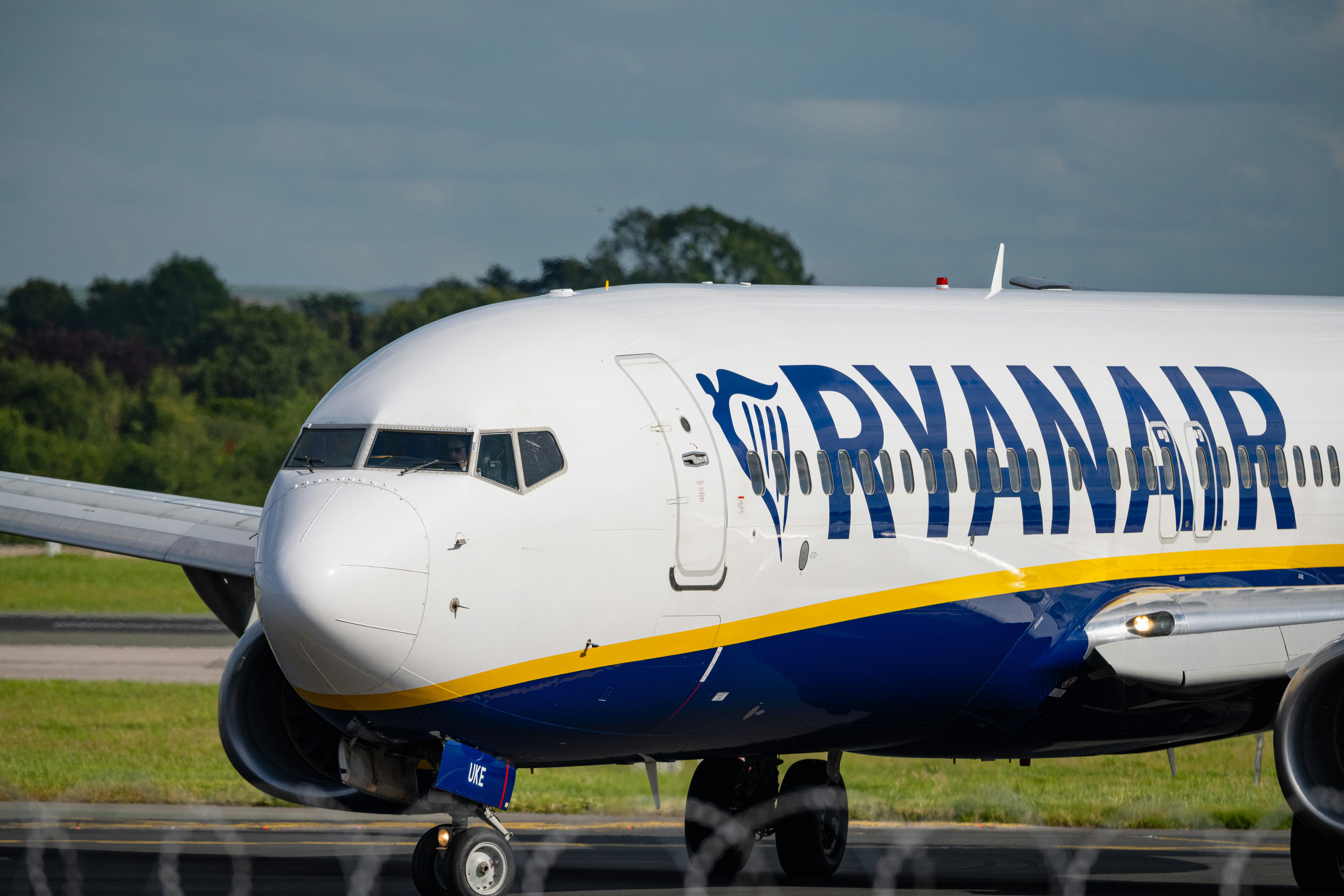 A Ryanair airplane is taxiing on a runway at an airport. The aircraft's nose and front fuselage are prominently visible, with the airline's name "RYANAIR" written in large blue letters on the side. The background shows a grassy area and trees under a cloudy sky.