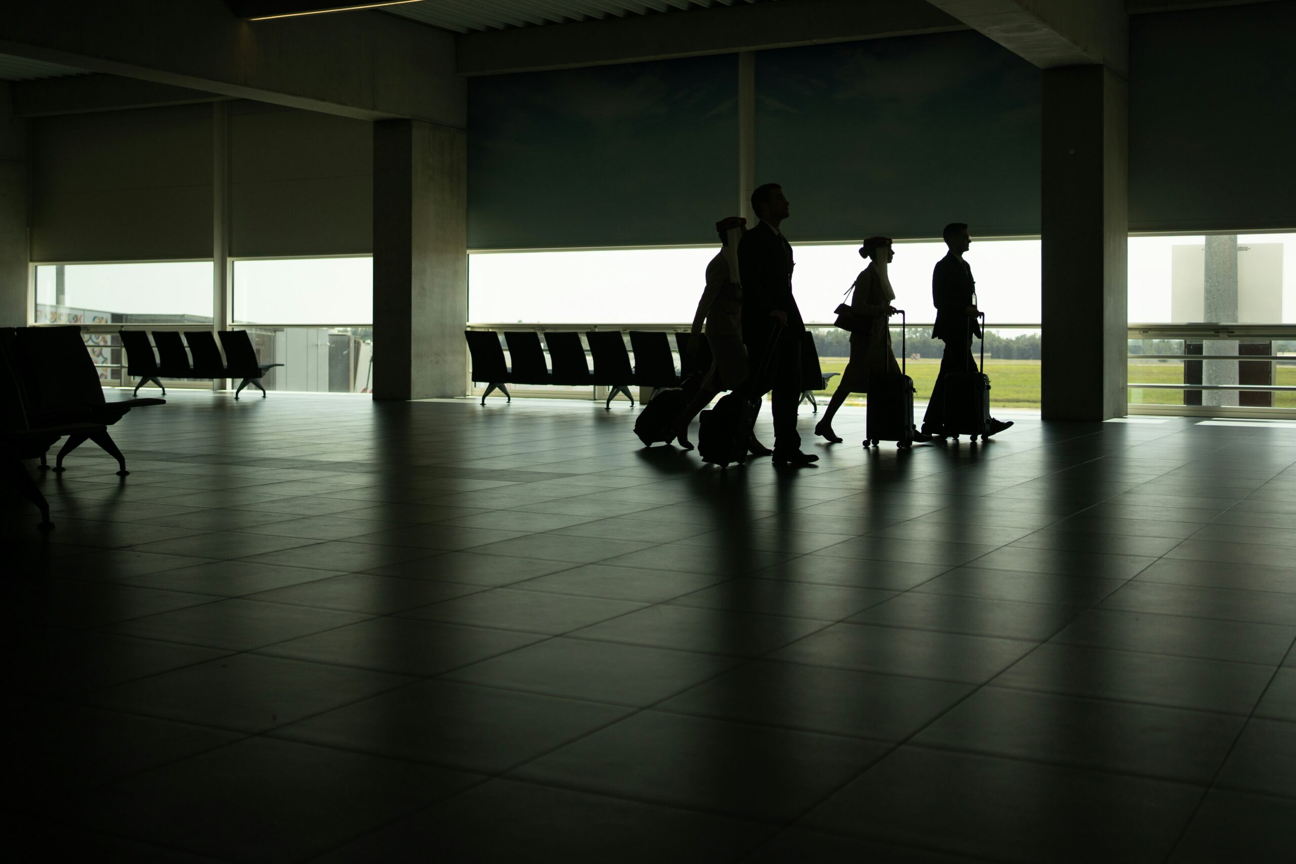 A silhouette of four people walking through an airport terminal, each pulling a wheeled suitcase. The terminal is spacious with large windows allowing natural light to illuminate the scene. Empty rows of seats are visible in the background.