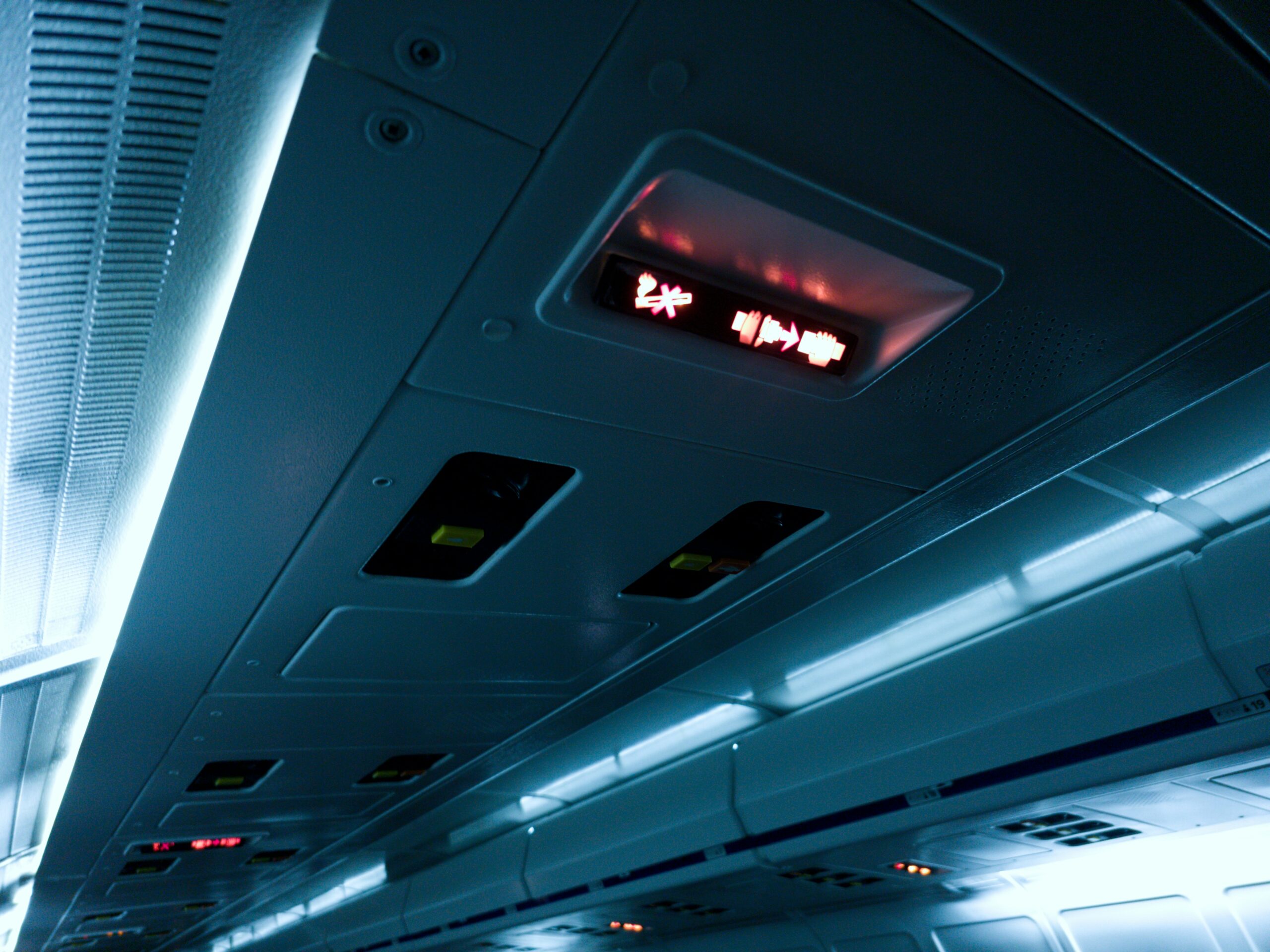 The interior ceiling of an airplane cabin showing illuminated signs. One sign displays a red "no smoking" symbol and a white "fasten seatbelt" symbol.
