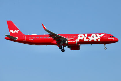 One of PLAY's bright red planes with white text (A321neo) in the air above Stansted airport.