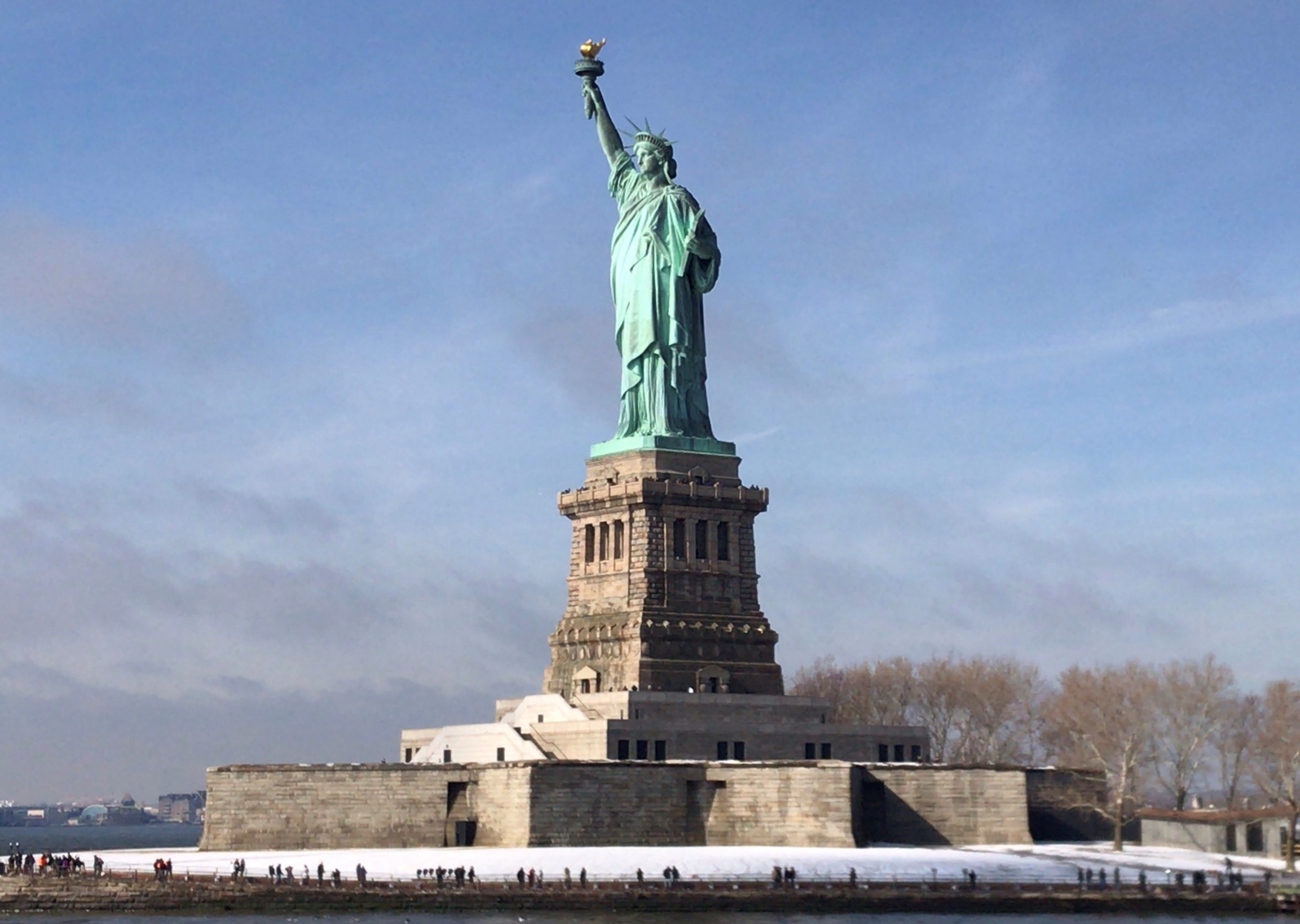 The Statue of Liberty in New York, USA during Winter. There is some snow around the base of the statue and trees with no leaves in the background.
