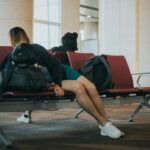 A person is lying down on a row of maroon airport seats, using a backpack as a pillow. The person is wearing a dark jacket, teal shorts, and white sneakers. Another person is seated in the background with their back turned to the camera.