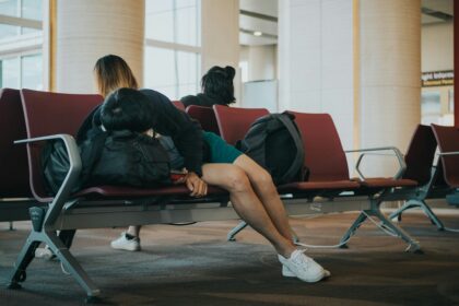 A person is lying down on a row of maroon airport seats, using a backpack as a pillow. The person is wearing a dark jacket, teal shorts, and white sneakers. Another person is seated in the background with their back turned to the camera.
