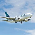 A WestJet aircraft is captured mid-flight against a backdrop of a partly cloudy blue sky. The airplane's landing gear is extended, indicating it is either approaching for landing or has just taken off.