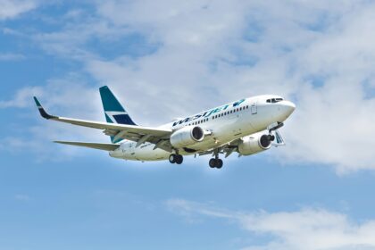 A WestJet aircraft is captured mid-flight against a backdrop of a partly cloudy blue sky. The airplane's landing gear is extended, indicating it is either approaching for landing or has just taken off.