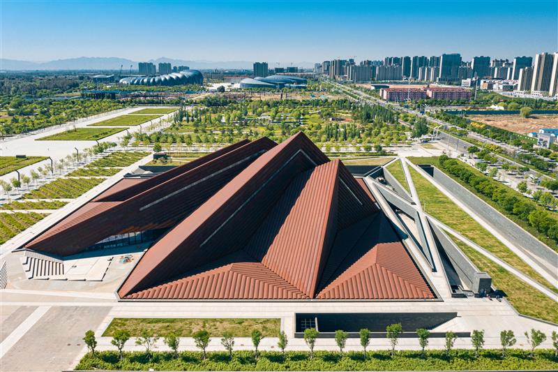 Aerial view of the Datong Art Museum, showcasing its unique angular and geometric architecture with rust-colored roofs. The museum is surrounded by lush greenery, pathways, and a modern urban landscape with high-rise buildings in the background.