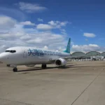 A Greater Bay Airlines aircraft is parked on the tarmac at an airport with a clear blue sky and scattered clouds in the background. The terminal building with its distinctive curved roof design is visible behind the plane.