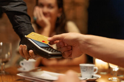 A close-up image shows a person holding a credit card near a payment terminal, ready to make a contactless payment. The card is gold-colored with the name "Chris Muller" visible on it. Another hand is holding the payment terminal. In the background, there are blurred figures of people sitting at a table with cups and glasses.