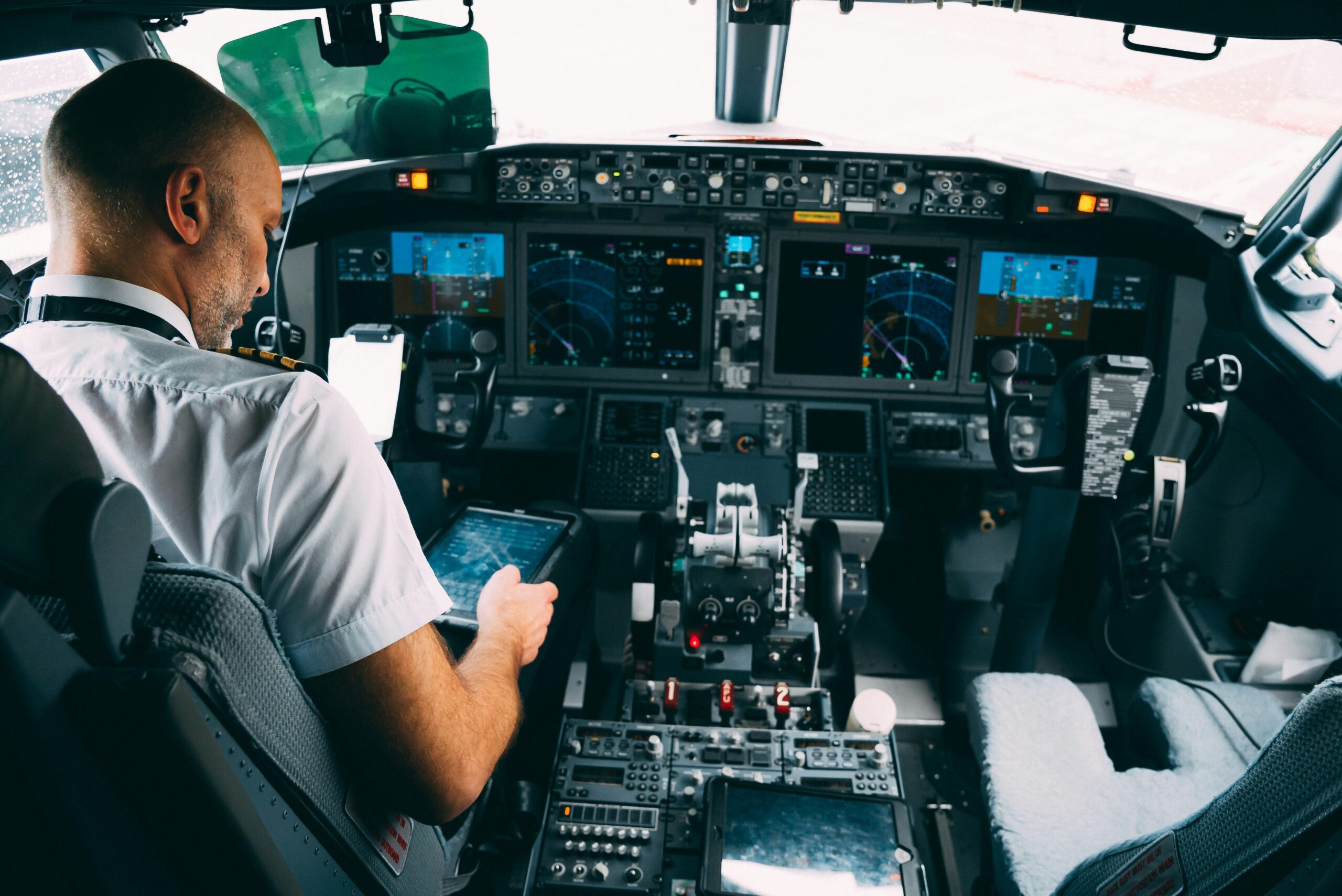 A pilot is seated in the cockpit of an aircraft, facing the control panel. The pilot's face is blurred for privacy. The cockpit is equipped with multiple screens displaying flight information, radar data, and navigation charts. The pilot holds a tablet device in one hand and appears to be interacting with it.