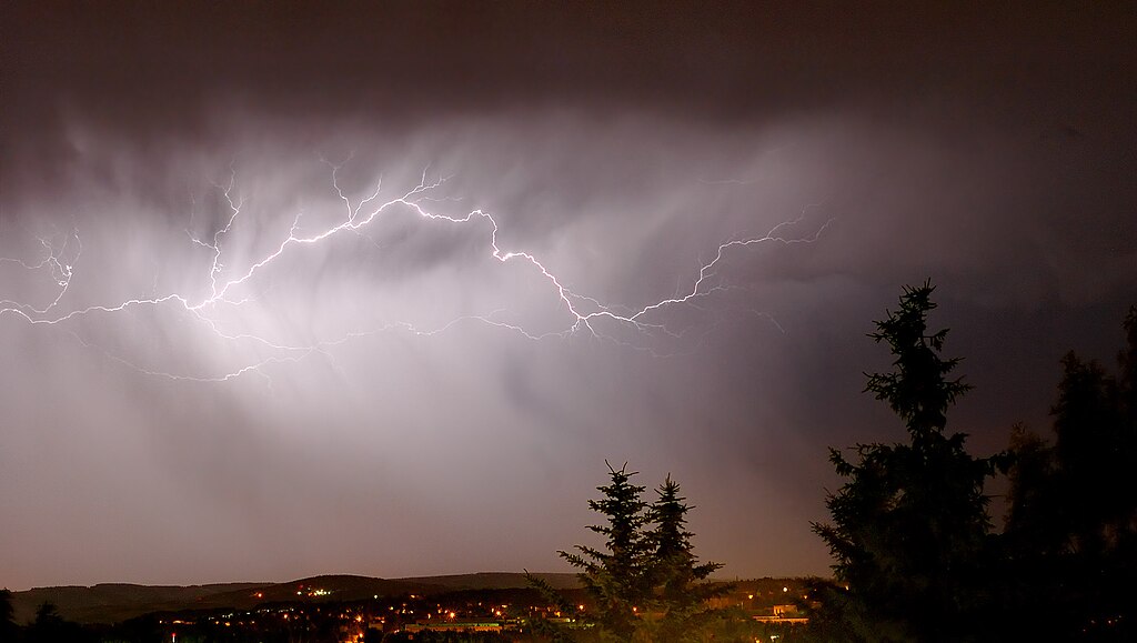 A vivid lightning bolt stretches across a dark, cloudy night sky above a distant town, illuminating the clouds and silhouetted trees in the foreground.