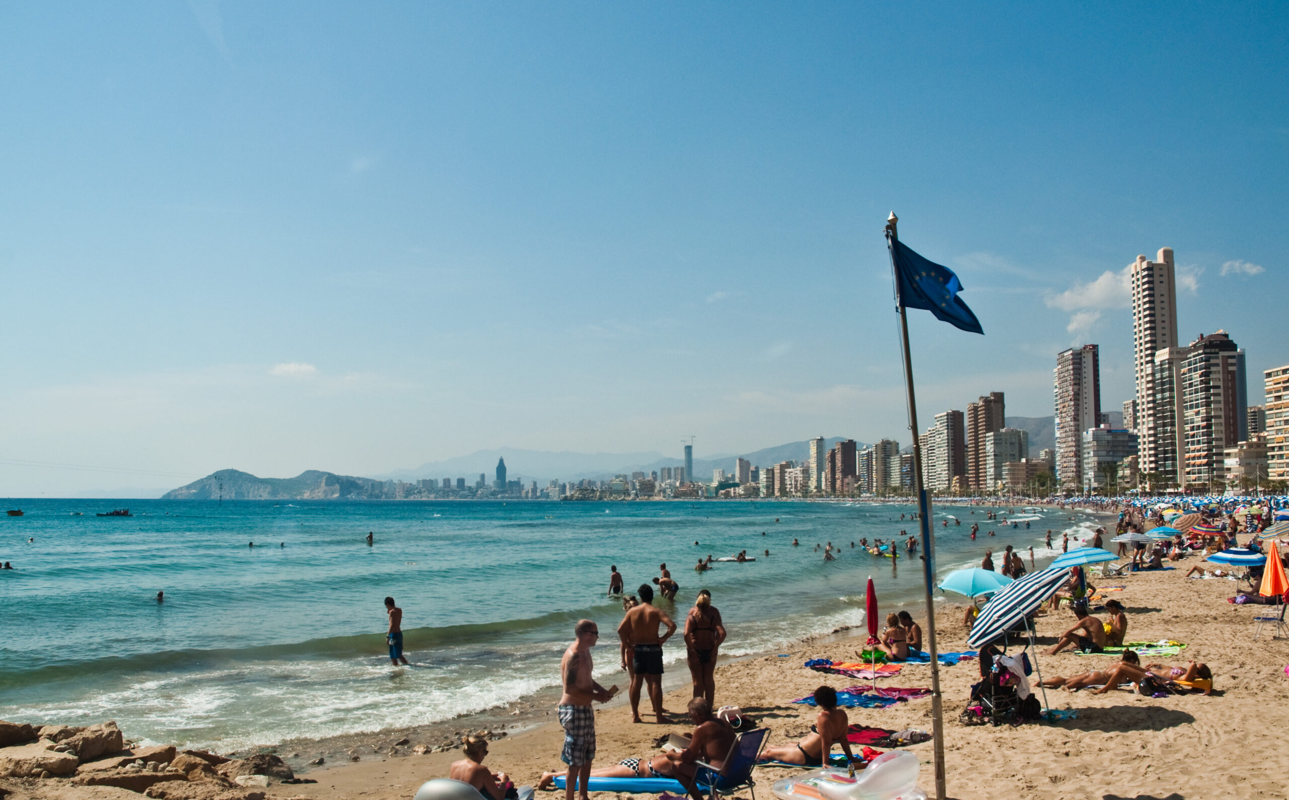 People lounging on a beach in the Costa Blanca with high-rise buildings visible in the background.