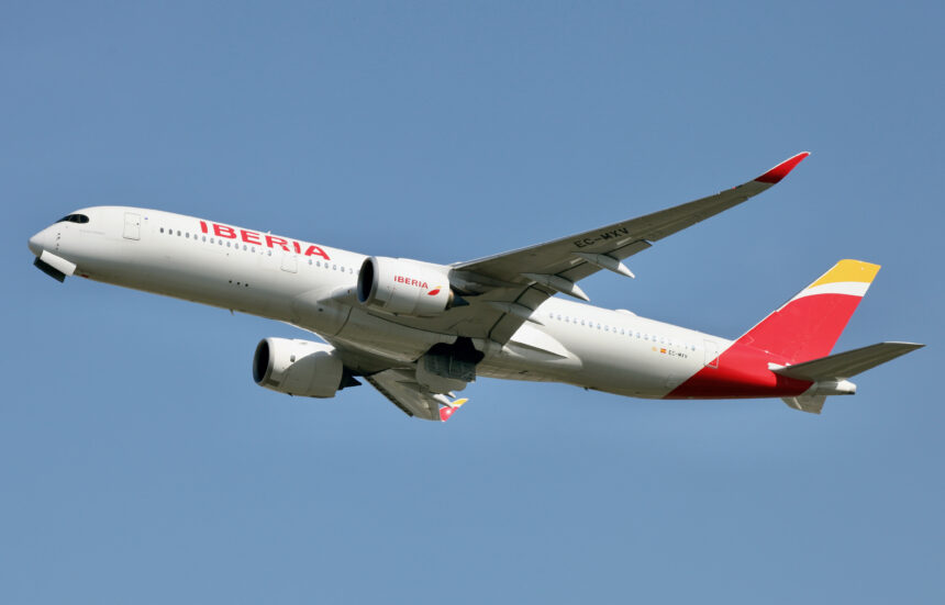 A white Iberia plane with a yellow and red tail flies through a blue sky.