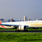 An Emirates plane lands on the tarmac with a large city skyline in the background filled with high-rise buildings.