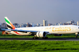 An Emirates plane lands on the tarmac with a large city skyline in the background filled with high-rise buildings.