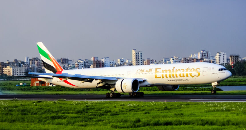 An Emirates plane lands on the tarmac with a large city skyline in the background filled with high-rise buildings.