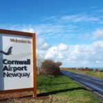 Cornwall Airport Newquay sign by the road, surrounded by grass. Blue sky with some white clouds in the background.