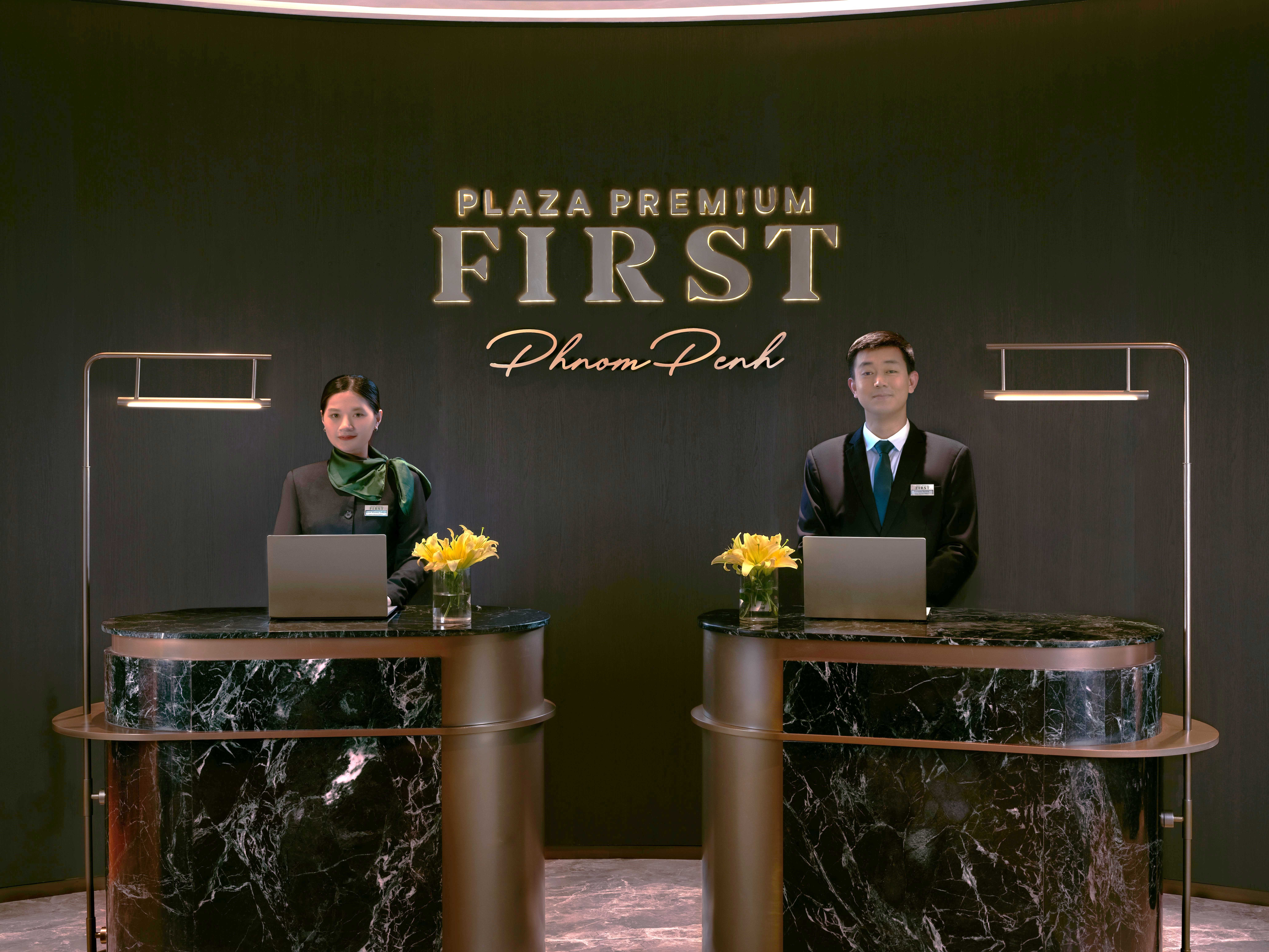 Two formally dressed attendants, one male and one female, wait at desks labelled Plaza Premoum First.