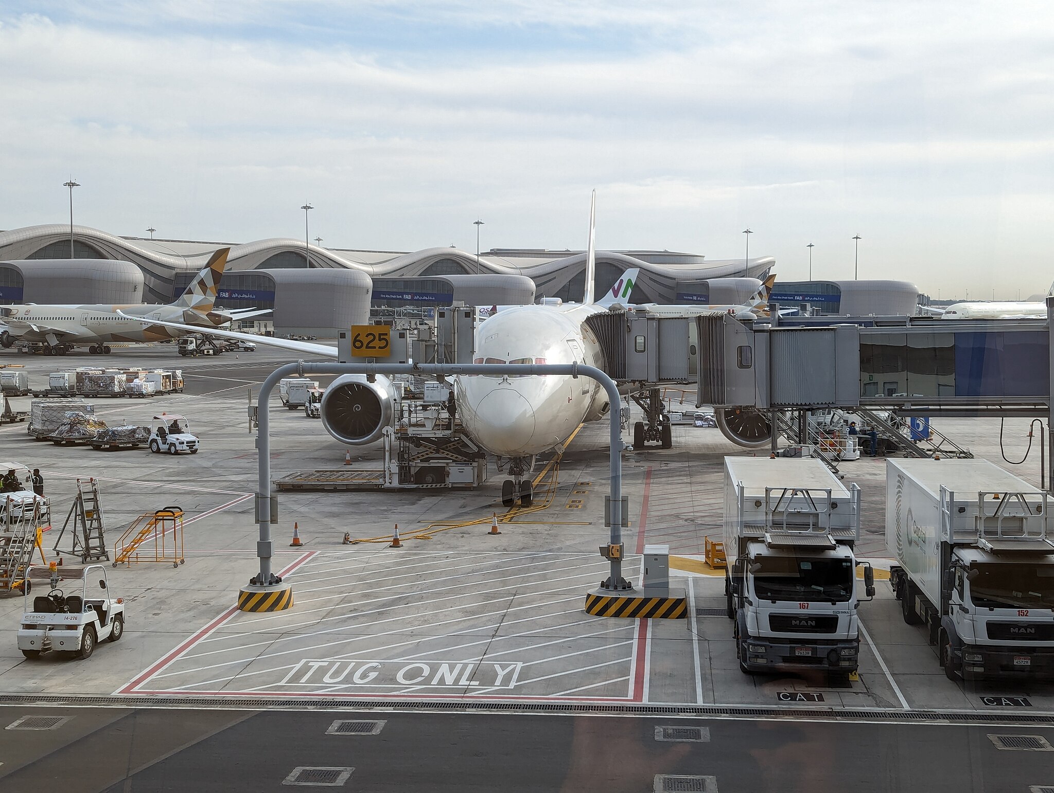Photo of a wide-body aircraft parked at an airport gate. It is connected from its left side to the terminal building via a passenger boarding bridge (aka air bridge). On its right side luggage is being loaded onto it. In the background are the other terminal buildings of Abu Dhabi Airport Terminal A.