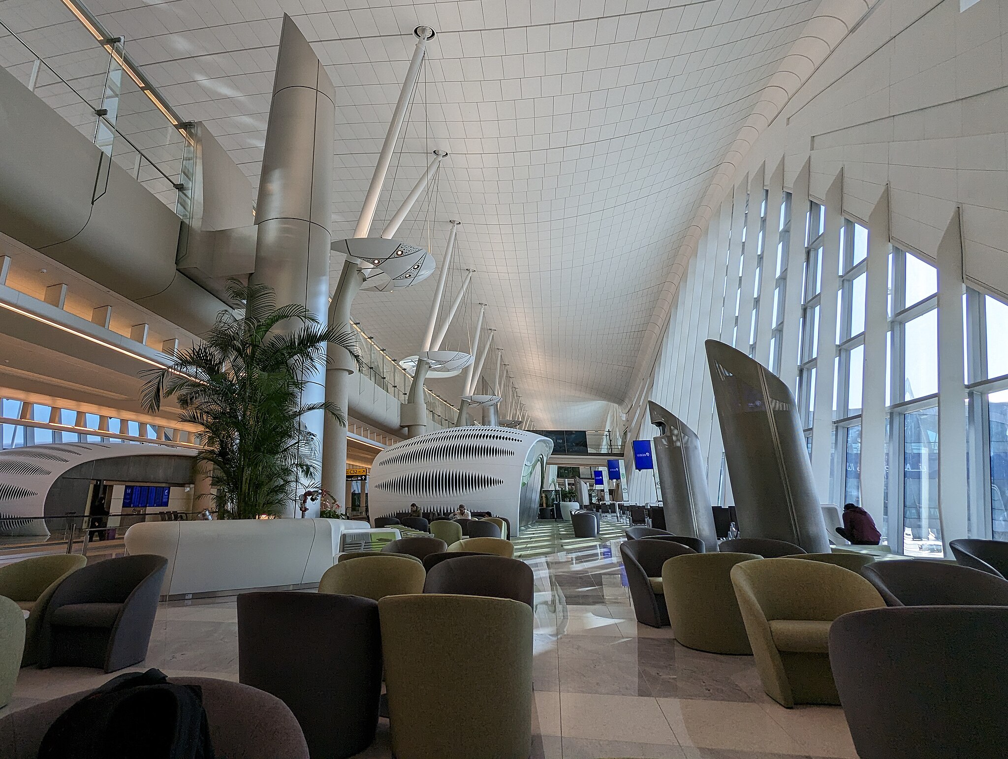 Photo of a large, heigh-ceiling, mostly empty airport departure gate. This section of the building is at least two storeys high, with a wide, elevated walkway running through the centre. In the departure are are several round-backed single-seater sofas for waiting passengers to sit on.