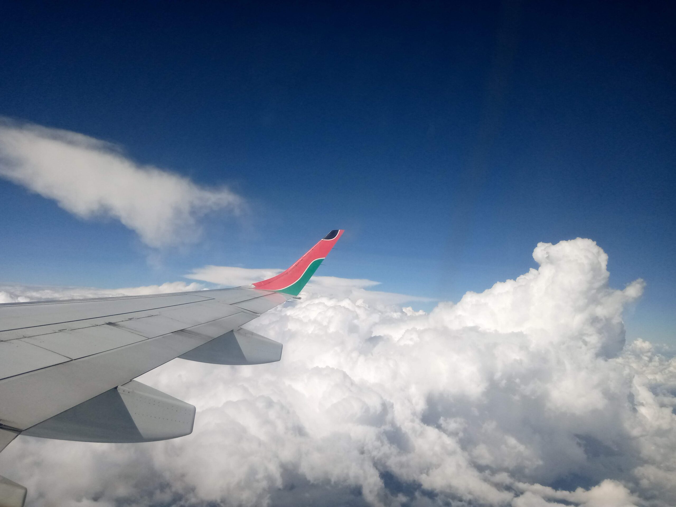 An aircraft wing with a red-and-green tip flies above bright white clouds and a deep blue sky, illustrating the growth of Uganda aviation, the Uganda ICAO Council 2025, Uganda international air service agreements 2025, ICAO Uganda, and Uganda aviation sector expansion ICAN2025.