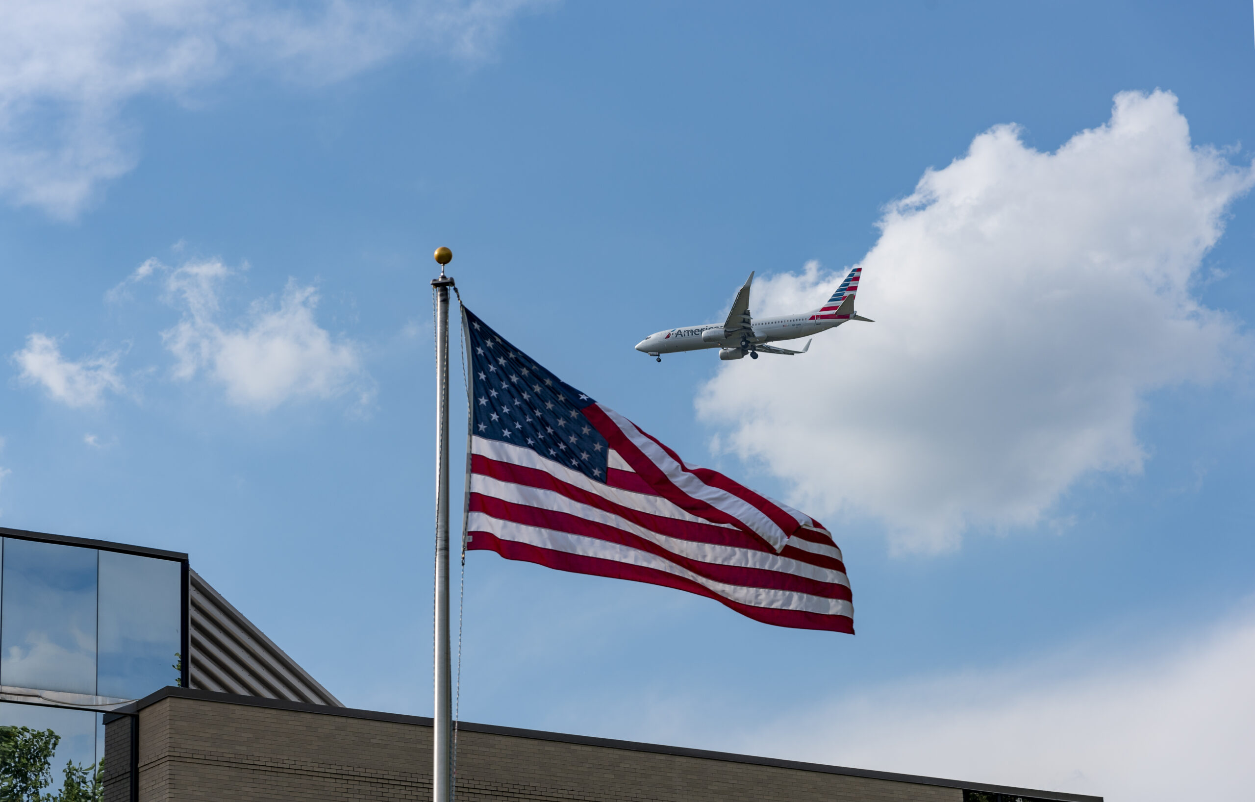 An American Airlines jet passes over the U.S. flag during approach, symbolising how the U.S. government shutdown and FAA flight cuts are impacting national air travel.