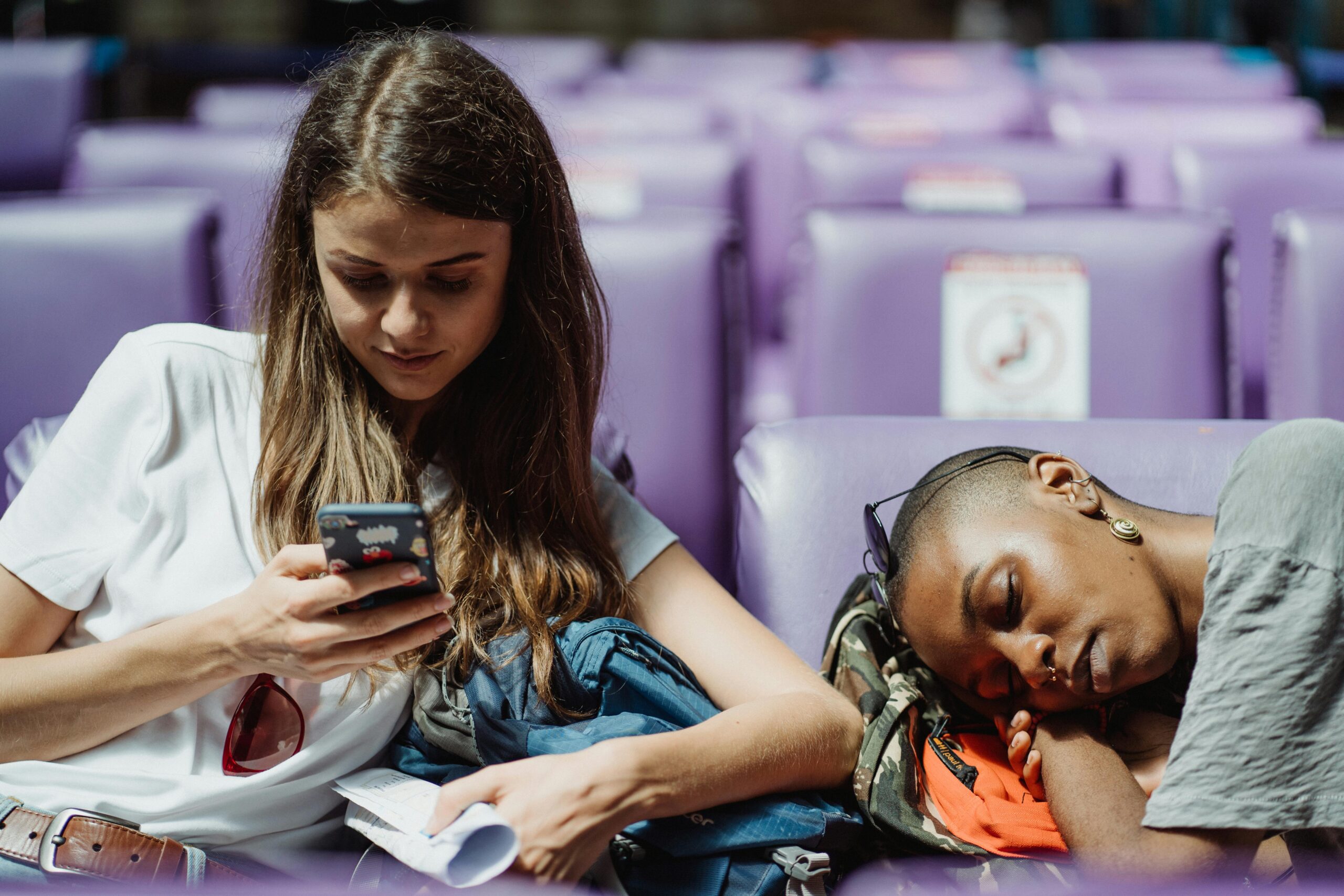 Women Waiting at the Station, one scrolling on the phone and other sleeping on a backpack