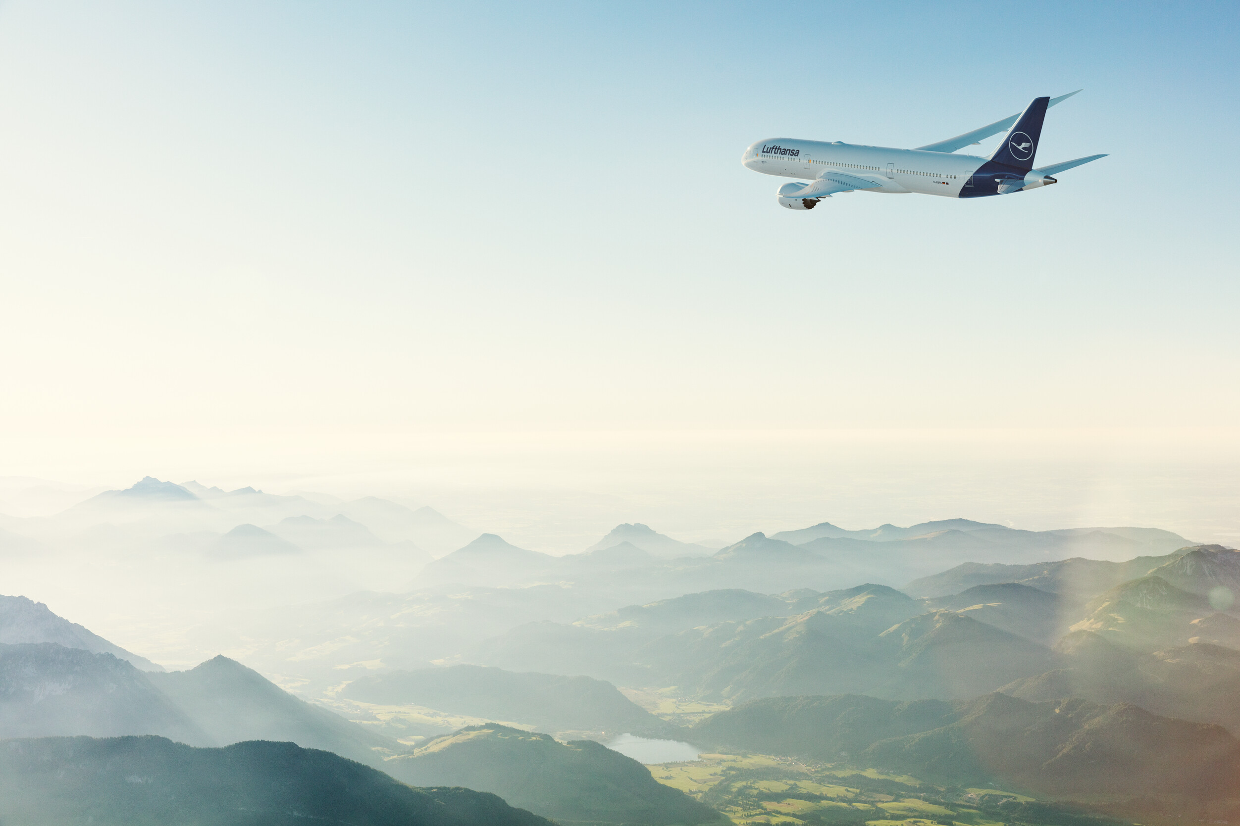 A Lufthansa plane flies high above mountains obscured by fog at dawn.