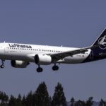 A white Lufthansa aircraft with blue tail and the Lufthansa crane logo visible, coming in for landing with landing gear out.