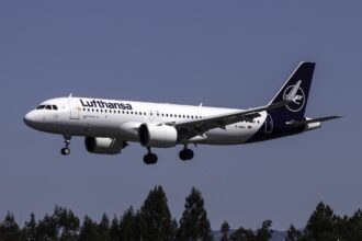 A white Lufthansa aircraft with blue tail and the Lufthansa crane logo visible, coming in for landing with landing gear out.