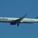 A JetBlue plane with landing gear out is high in a bright blue sky.