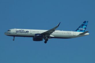 A JetBlue plane with landing gear out is high in a bright blue sky.