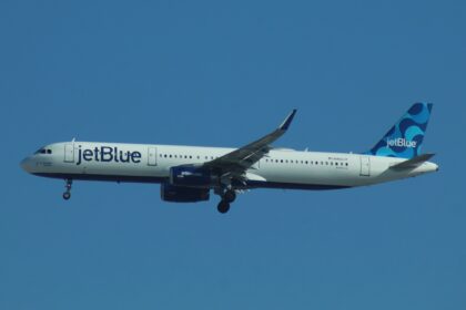 A JetBlue plane with landing gear out is high in a bright blue sky.