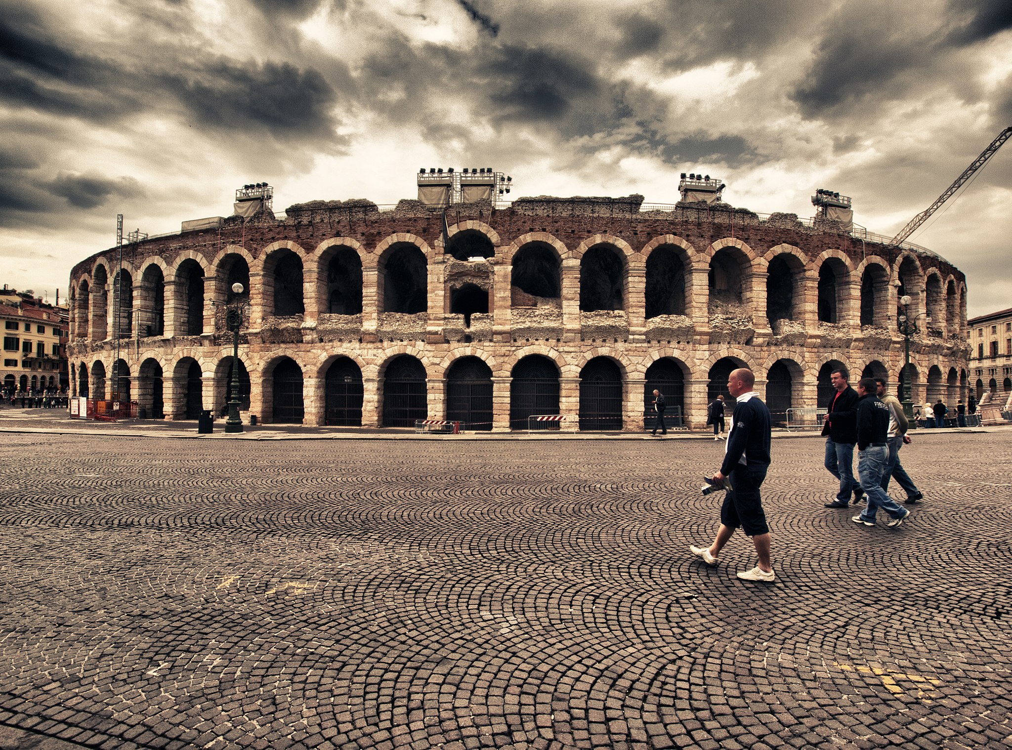 The Arena di Verona, a Roman theatre, is in the centre, with medieval paving around it and people walking past. The sky is cloudy and dramatic.