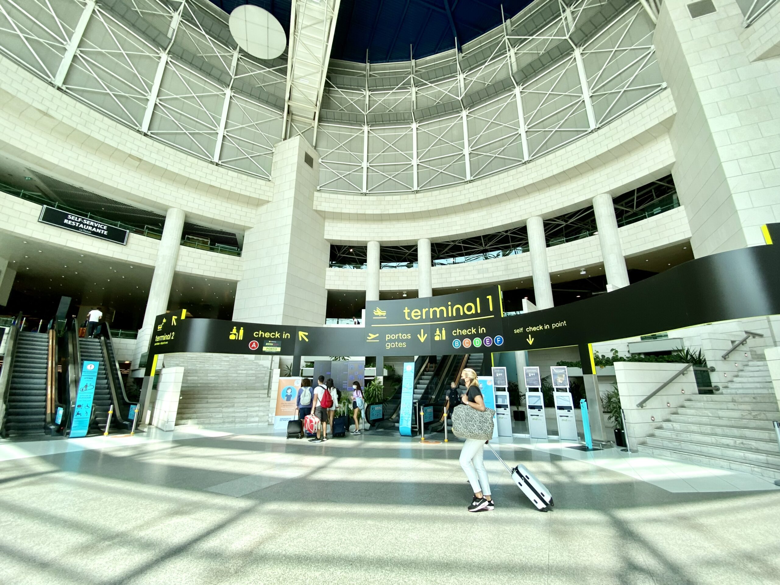 A sign reading "terminal 1" and others with directions hang over escalators and stairs inside the Lisbon Airport. Some people with suitcases can be seen passing by.
