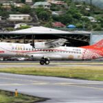 A side profile of an Air Tahiti ATR 72-600 turboprop aircraft landing on a runway. The plane features a special white and red livery with "60 ANS YEARS" gold lettering on the fuselage and intricate Polynesian-inspired patterns on the tail. A lush green hillside dotted with residential houses is visible in the background under an overcast sky.