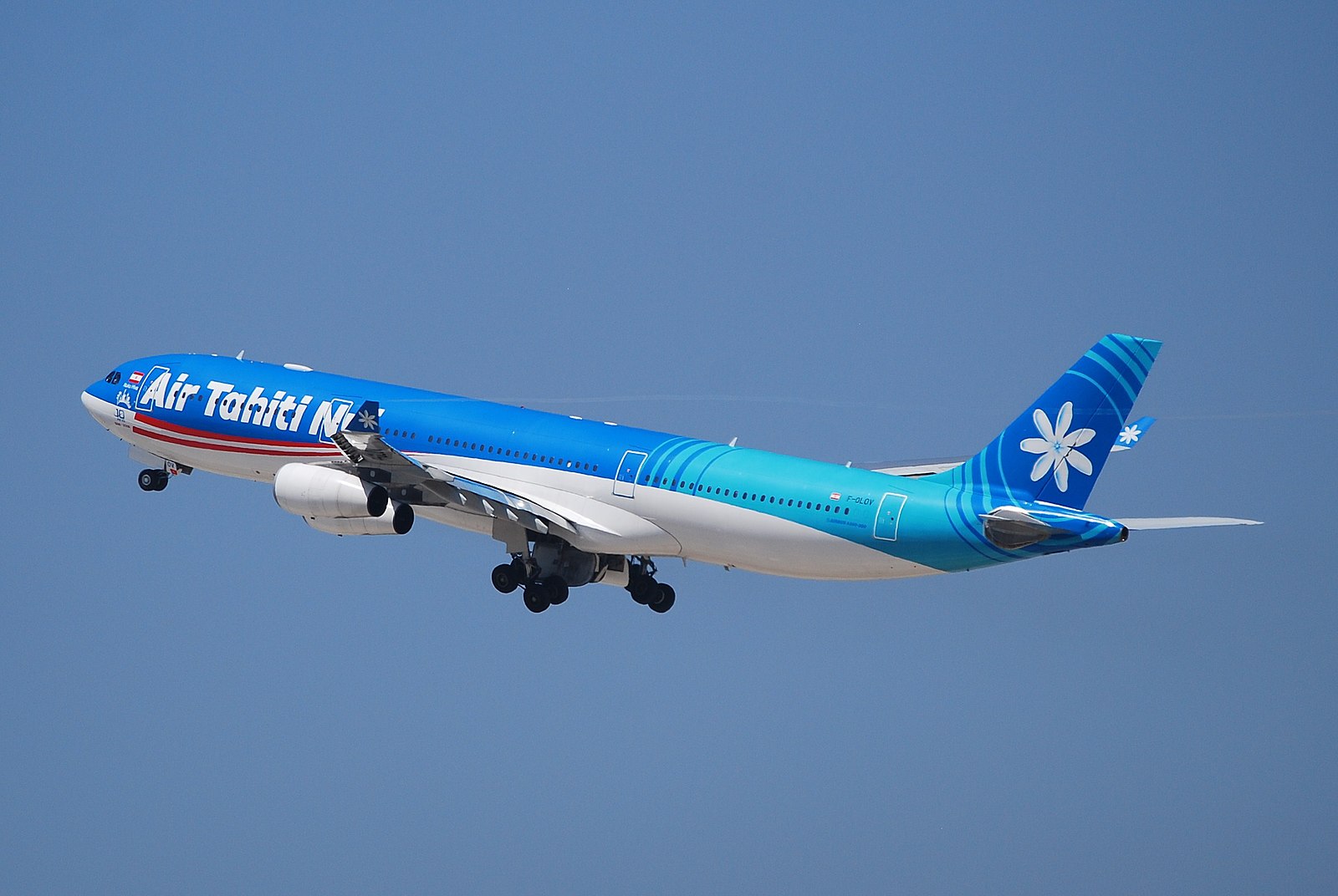 An Air Tahiti Nui Airbus A340-300 climbing into a clear blue sky. The aircraft features a vibrant blue and white livery with a red stripe along the forward fuselage. The tail is decorated with a large white Tiare flower (Tahitian gardenia) on a blue background with wave-like patterns. The landing gear is still deployed as the four-engine plane ascends.