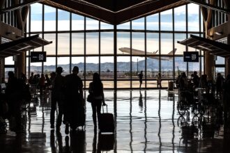 A high-contrast, wide-angle silhouette of an airport terminal interior. In the foreground, travelers with luggage and people in wheelchairs are silhouetted against a massive floor-to-ceiling window. The polished floor reflects the light and the figures perfectly. Through the glass, a large airplane is seen taking off against a backdrop of distant mountains and a clear sky. Signs for "Gates D6/D8" and "Gates D5/D7" hang from the ceiling on the left and right.