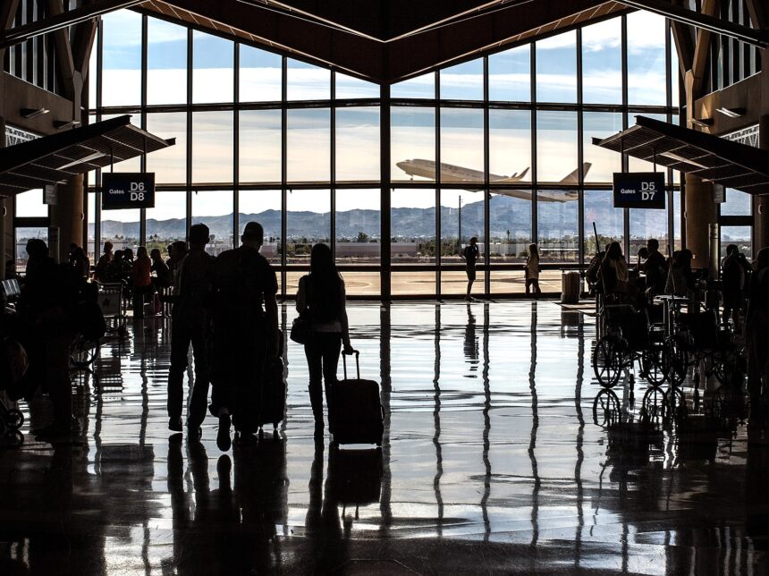 A high-contrast, wide-angle silhouette of an airport terminal interior. In the foreground, travelers with luggage and people in wheelchairs are silhouetted against a massive floor-to-ceiling window. The polished floor reflects the light and the figures perfectly. Through the glass, a large airplane is seen taking off against a backdrop of distant mountains and a clear sky. Signs for "Gates D6/D8" and "Gates D5/D7" hang from the ceiling on the left and right.