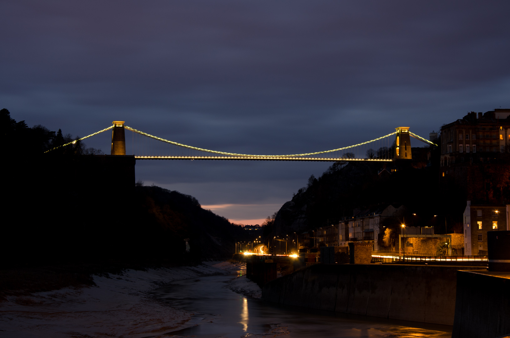 The iconic Clifton Suspension Bridge at dusk, lit up with houses in front of it at the bottom of the gorge.
