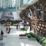 The interior of Indira Gandhi International Airport in Delhi, showing the iconic "Mudra" hand sculptures mounted on a wall of copper-colored discs. The terminal is bright and spacious with travelers walking through the concourse. A large advertisement for the Samsung Galaxy S24 Ultra is visible in the background.