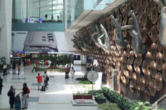 The interior of Indira Gandhi International Airport in Delhi, showing the iconic "Mudra" hand sculptures mounted on a wall of copper-colored discs. The terminal is bright and spacious with travelers walking through the concourse. A large advertisement for the Samsung Galaxy S24 Ultra is visible in the background.