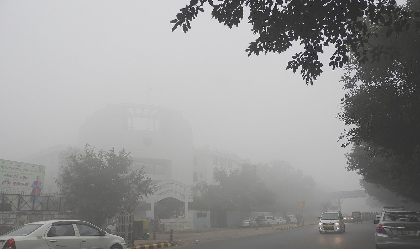 A hazy outdoor scene in Delhi showing a wide road filled with vehicles, including several white cars and motorcycles, under a thick layer of grey smog. Visibility is severely limited by the air pollution, and the silhouettes of trees and distant structures are barely visible through the dense haze.