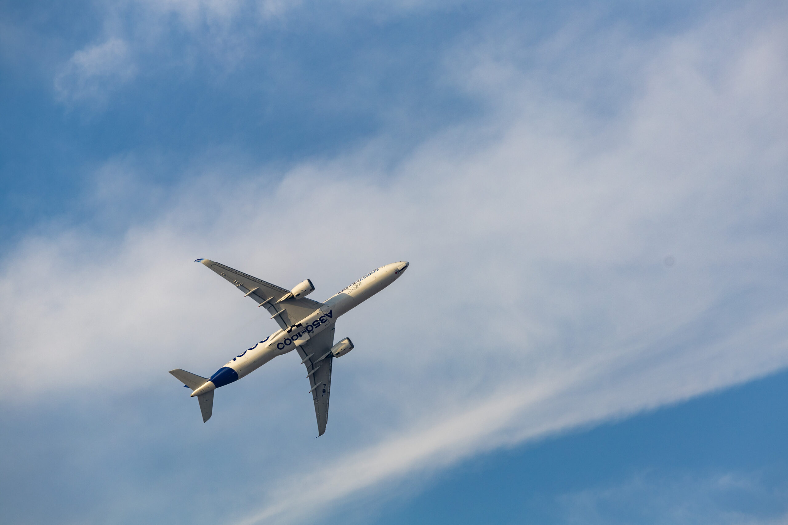 A white Airbus A350-1000 wide-body airplane, viewed from below, ascends into a blue sky with white clouds.