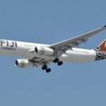 A wide-body Fiji Airways Airbus A330-200 (registration DQ-FJU) is captured from below against a clear blue sky as it prepares for landing. The aircraft features a distinctive white livery with black traditional Masi motifs on the engines and tail. The tail also includes a prominent circular brown design. The landing gear is fully deployed, and the "FIJI AIRWAYS" wordmark is clearly visible on the forward fuselage.