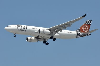 A wide-body Fiji Airways Airbus A330-200 (registration DQ-FJU) is captured from below against a clear blue sky as it prepares for landing. The aircraft features a distinctive white livery with black traditional Masi motifs on the engines and tail. The tail also includes a prominent circular brown design. The landing gear is fully deployed, and the "FIJI AIRWAYS" wordmark is clearly visible on the forward fuselage.